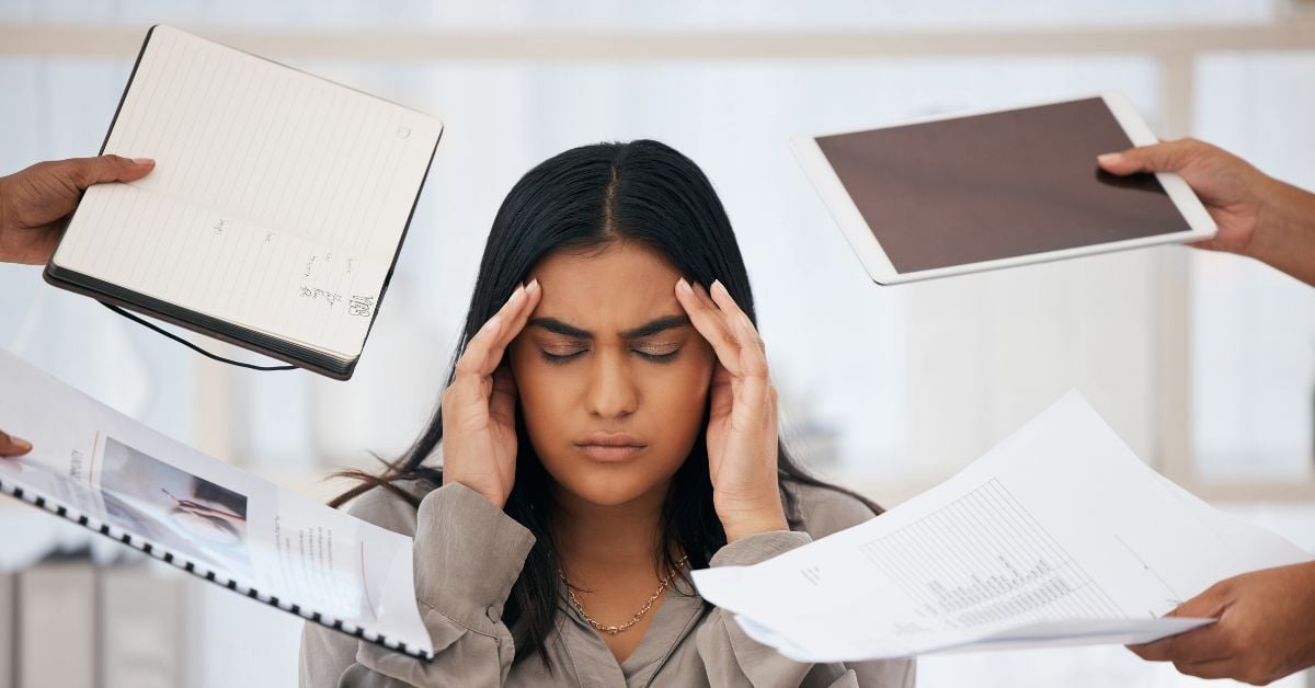 A woman is wondering how to deal with employee burnout at work as she holds her head and people hand her documents.